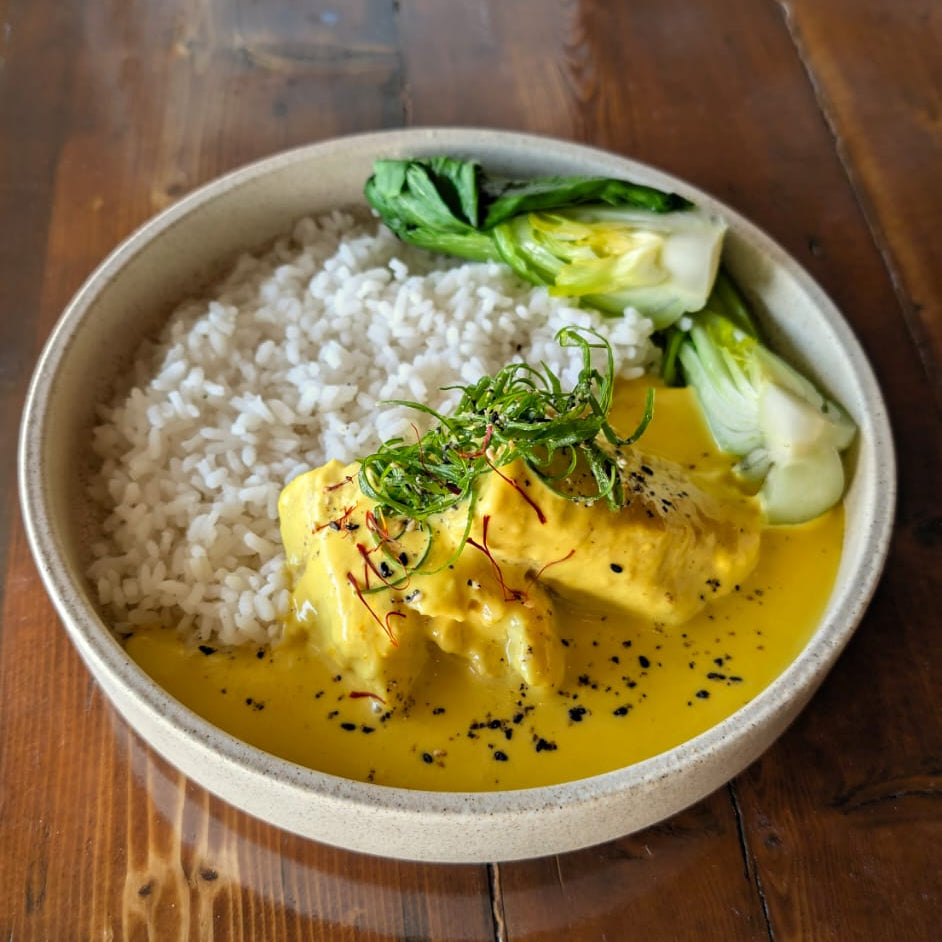 Bowl of saffron & coconut cod with yellow sauce, rice, and greens on a wooden table
